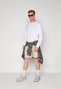 Fehér hosszú ujjú ing logóval, bézs rövidnadrág, szürke és sárga kockás ing a derék körül megkötve, napszemüveg és szürke sportcipő.