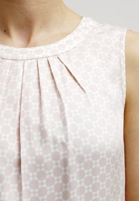 Hihaton vaaleanpunainen paita, jossa on hienovarainen geometrinen kuvio ja laskoksia pyöreän pääntien alapuolella henkilön ylävartalossa.