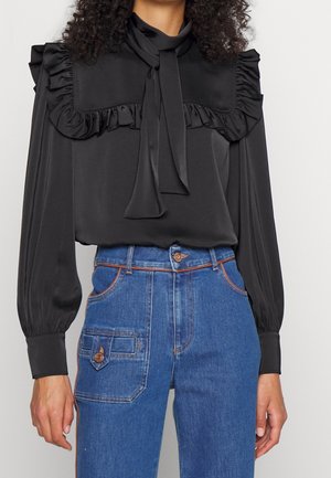 Kobieta w czarnej bluzce z falbanianym karczkiem i długimi rękawami, w połączeniu z wysokim stanem niebieskich dżinsów z boczną kieszenią.