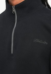 Fekete zipzáros fleece pulóver magas gallérral, ezüst színű cipzárral és diszkrét hímzett "Hollister" logóval a mellkasán. Puha tapintású.
