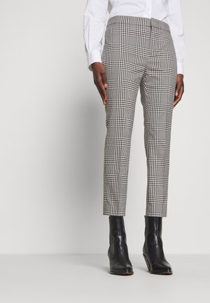Personne portant un pantalon slim à carreaux noir et blanc, des bottines noires et une chemise blanche à manches longues, debout devant un fond uni.