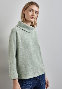 Ung kvinna med ljust hår som bär silvriga hoops örhängen, en ljusgrå polotröja och mörkblå jeans mot en neutral bakgrund.