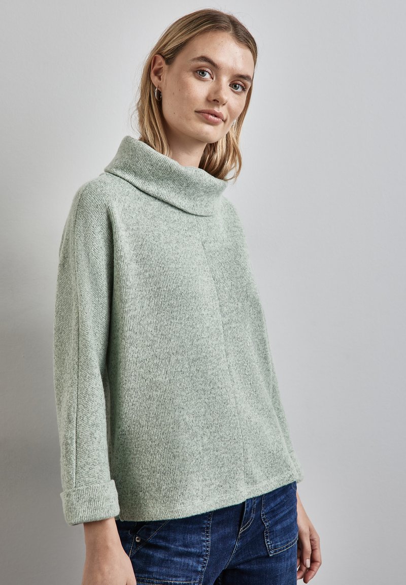 Ung kvinna med ljust hår som bär silvriga hoops örhängen, en ljusgrå polotröja och mörkblå jeans mot en neutral bakgrund.