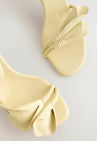 Paire de sandales plates jaune pâle avec des brides torsadées et des détails en cuir en forme de pétales, présentées sur une surface claire.