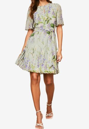 Vrouw draagt een lichtgroene bloemenjurk met geplooide korte mouwen, een taillekoord en witte sandalen met hoge hakken en bandjes.
