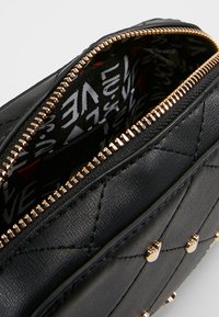 Musta tikattu synteettisestä nahasta valmistettu laukku, jossa kullanvärinen vetoketju, kuvioitu kangasvuori ja koristeelliset niitit ulkopuolella.