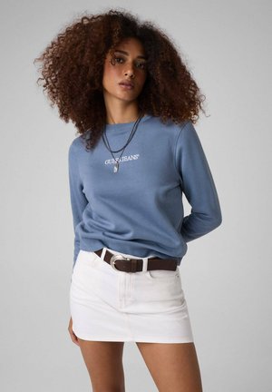 Donna con capelli ricci che indossa una felpa blu Guess Jeans e una gonna bianca con cintura marrone, in piedi contro uno sfondo semplice.
