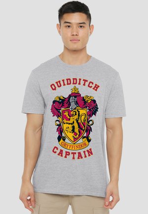 Young man wearing grey t-shirt with red and yellow Gryffindor Quidditch Captain crest and beige pants.