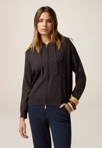 Hoodie noir zippé en tissu doux, doté d'embellissements dispersés, d'une capuche avec cordons de serrage et de poignets côtelés. Porté avec un pantalon marine.