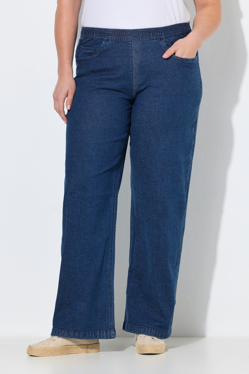 Personne portant un jean bleu foncé à jambes larges avec une taille élastique, une main dans la poche, associée à des chaussures beiges, debout sur fond blanc.