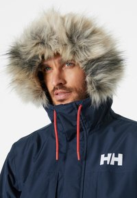 Parka impermeable de color navy con capucha forrada de pelo sintético, cordones rojos y un logo blanco en el pecho. Presenta un cuello alto y cierre con cremallera.