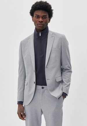 Lichtgrijze gestreepte colbert met bijpassende broek, gedragen boven een aansluitend donkerblauwe ritsvest. Glad stof, op maat gesneden, subtiele textuur.