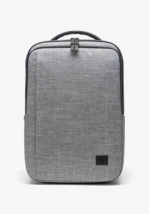 Sac à dos en tissu gris rectangulaire avec tirettes de fermeture éclair noires et une poignée noire, affichant une petite étiquette de marque noire en bas à droite.