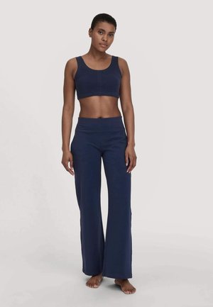 Frau in einem dunkelblauen ärmellosen Crop-Top und weiten Hosen, barfuß vor einem schlichten hellen Hintergrund stehend.