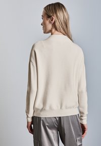 Maglione lavorato a maglia beige chiaro con scollo rotondo, spalle basse e orlo a coste. Indossato con pantaloni in seta grigi lucenti. Texture liscia.