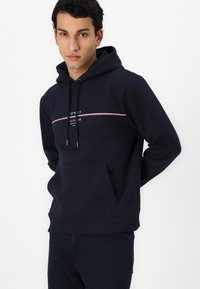 Navy blue hoodie made of soft cotton with a front kangaroo pocket, featuring a white and red logo strip across the chest.