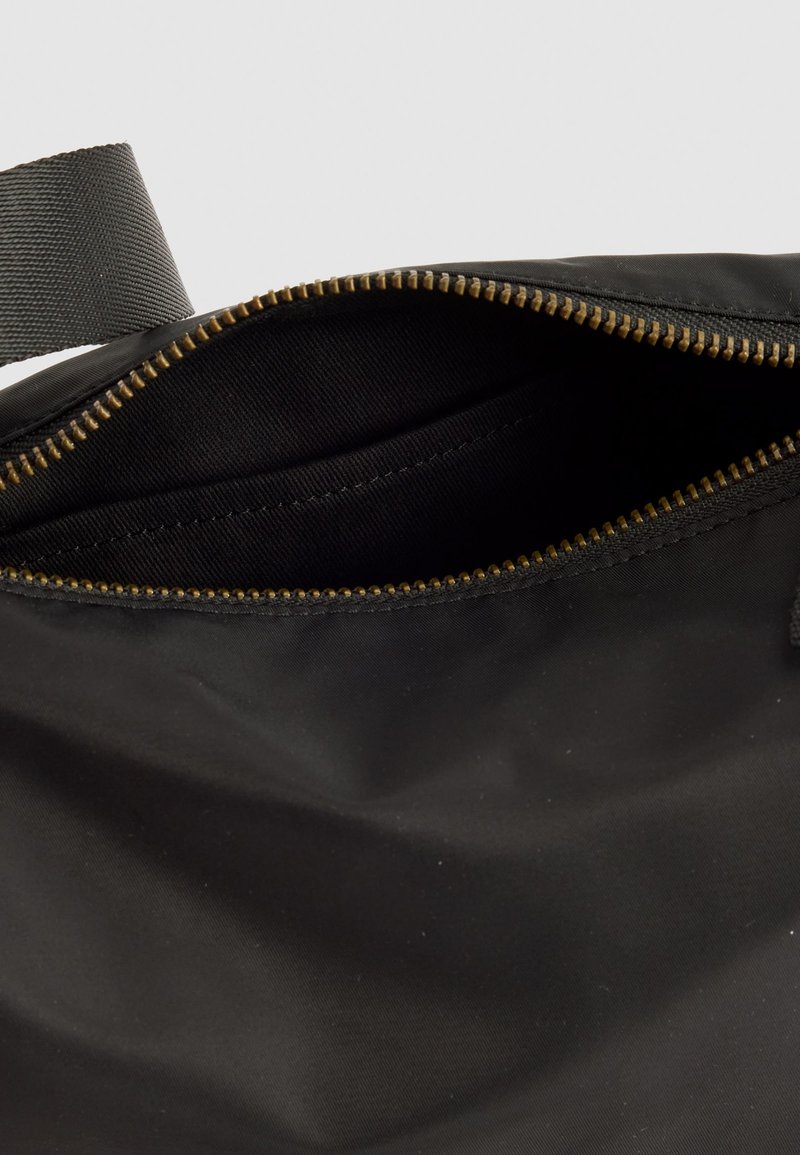 Bolsa de tela negra parcialmente abierta con un visible cierre dorado y una cinta negra contra un fondo claro.