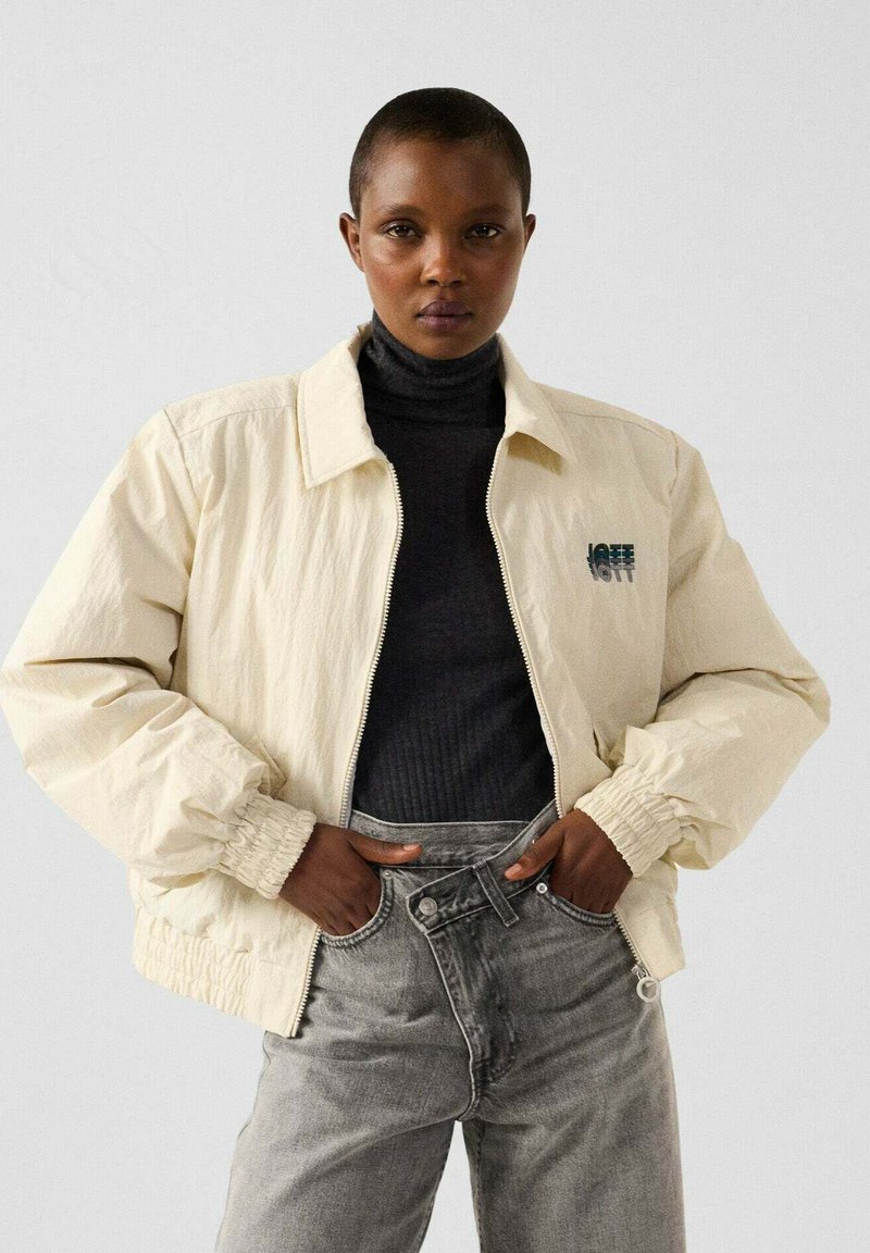 Giacca bomber di colore crema chiaro con polsini elasticizzati, chiusura con zip e dettaglio del logo; abbinata a un dolcevita scuro e jeans grigi a vita alta.