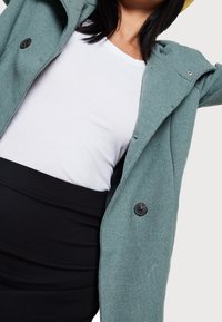 Groene getextureerde jas met grote knopen, gedragen over een witte geribde top en een zwarte aansluitende rok. De jas heeft een gestructureerde kraag en een open voorkant.