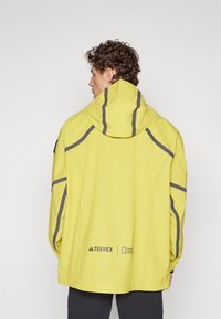 Κίτρινο αδιάβροχο μπουφάν με γκρι λεπτομέρειες και ανακλαστική επένδυση. Διαθέτει κουκούλα και χαλαρή εφαρμογή. Μάρκα στο κάτω μέρος της πλάτης.