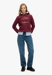 Sudadera color burdeos con bolsillo frontal y cordón, adornada con texto dorado, superpuesta sobre una camisa de botones azul claro. Combinada con jeans azules y zapatos negros.