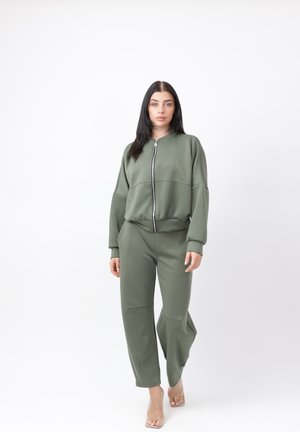 Donna con capelli lunghi e scuri che indossa una giacca verde oliva con zip e pantaloni abbinati, che cammina in avanti su uno sfondo bianco semplice.