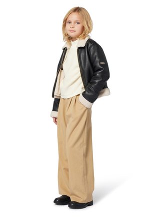 Enfant portant une veste en shearling noir, un pull en tricot crème, un pantalon beige large et des chaussures noires, se tenant les mains dans les poches.