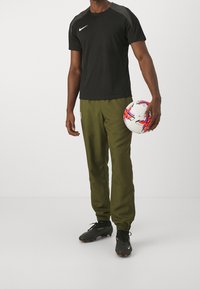 Černé sportovní tričko Nike s krátkým rukávem, olivově zelené kalhoty s elastickými manžetami a černé kopačky, držící bílý fotbal.