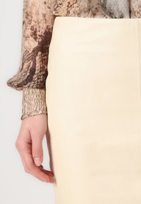 Lærpencilskjørt i tan med jevn tekstur, kombinert med en gjennomsiktig, mønstrete bluse med slangeskrift i nyanser av brunt og beige.