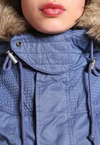 Blå kviltad jacka med en pälsfodrad huva, dragband och metallbeslag. Slät textur och en lager på lager-kragsdesign.