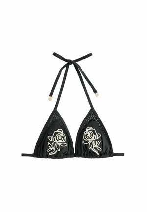 Femeie cu părul udat, purtând un bikini negru cu broderie de trandafiri albi, stând pe o plajă stâncoasă lângă două șezlonguri din lemn cu pernuțe albe.