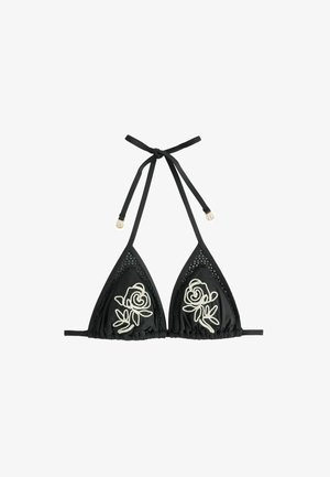 Femeie cu părul udat, purtând un bikini negru cu broderie de trandafiri albi, stând pe o plajă stâncoasă lângă două șezlonguri din lemn cu pernuțe albe.
