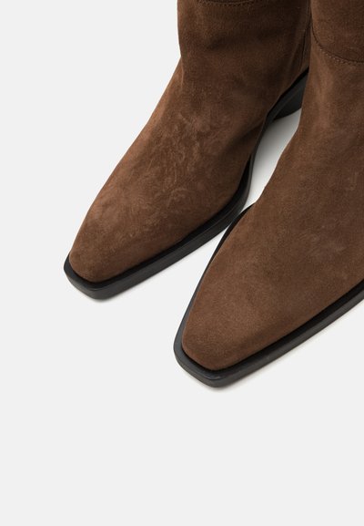 Braune Wildleder-Ankelstiefel mit spitzem Zehendesign, ausgestattet mit einer flachen schwarzen Sohle und glatter Textur. Keine sichtbaren Verzierungen oder Muster.