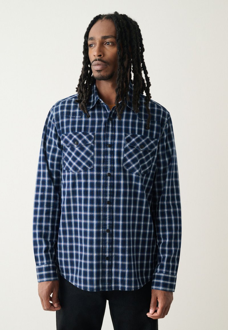 Chemise en flanelle à carreaux bleu et blanc avec deux poches poitrine, boutonnage sur le devant, manches longues et ourlet incurvé. Texture douce.