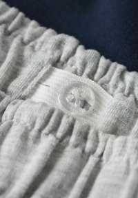 Grå tygmidja med rynkad textur, med en genomskinlig plastknapp och en vit flik för knäppning. Bakgrunden är mörkblå.