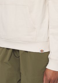 Felpa beige chiaro con tasca frontale, polsini e orlo a coste. Presenta una piccola toppa con logo in basso a sinistra. Indossata con pantaloni verde oliva.