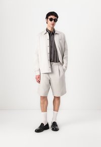 Lysegrå linnedjakke, rutet skjorte og matchende shorts, parret med sorte loafers og hvide sokker. Tilbehør inkluderer mørke solbriller.