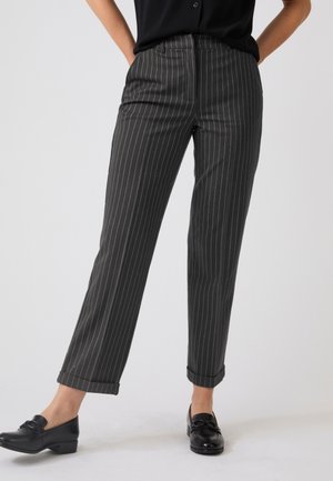 Persona che indossa pantaloni neri a righe sottili con orli risvoltati e mocassini neri in pelle, in piedi davanti a uno sfondo semplice.