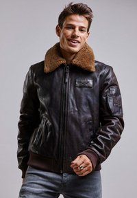Veste en cuir marron de style aviateur avec un col en shearling beige, fermeture éclair frontale et poches latérales. Présente un patch en tissu sur la poitrine.