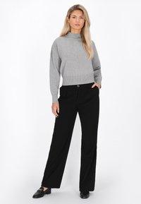 Eine Frau steht bekleidet mit einem grauen Langarmpullover, schwarzen weiten Hosen und schwarzen Loafers vor einem weißen Hintergrund.