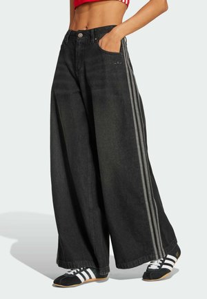 Personne portant un pantalon noir à jambes larges avec des bandes latérales et des baskets noires à rayures blanches, debout avec une main dans la poche.