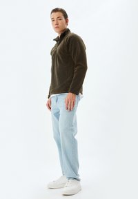 Chaqueta de forro polar verde oliva con un alto cuello, cierre de cremallera en la parte delantera, combinada con jeans de un azul claro y zapatillas blancas. Textura suave y ajuste relajado.