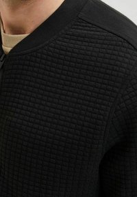 Černá strukturovaná mikina s vyvýšeným mřížkovým vzorem, kulatým výstřihem a kontrastním světlým materiálem uvnitř. Přední zapínání na zip.