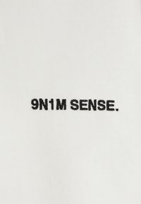 Fehér, bordázott anyag fekete hímzett szöveggel, amely így olvasható: "9N1M SENSE." A szöveg középen helyezkedik el, és kiemelkedik a háttérből.
