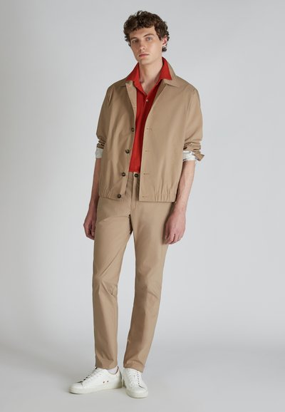 Giacca leggera beige con bottoni sopra una camicia rossa con colletto, abbinata a pantaloni beige e sneakers bianche. Tessuto liscio, vestibilità comoda.