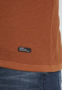 Pulover tricotat de culoare ruginie, cu țesătură texturată. Are o etichetă neagră cu text alb cusut pe margine. Design țesut și manșete cu nervuri.