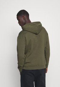 Olivengrønn hettegenser med kengurulomme, myk tekstur, oversized passform og snor i hetta, sett fra baksiden.