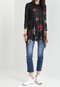 Tunique noire avec des motifs floraux rouges et blancs, associée à un jean bleu avec des détails brodés, et des chaussures de sport blanches.