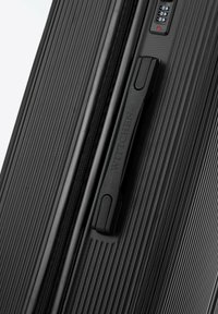 Black suitcase with a textured exterior, featuring horizontal grooves, a black zipper, and a molded handle with branding. Lock mechanism visible.