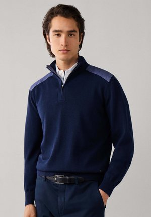 Joven con un jersey azul marino con cremallera hasta el pecho, con parches azul claro en los hombros y pantalones oscuros con cinturón negro, de pie frente a un fondo liso.